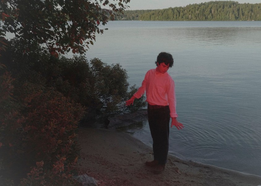 A Man wearing a white long sleeve top, black trousers, brown belt, and brown shows, with curly dark brown hair stands at the edge of a lake on the sand with his back to the water. There is a red hued light shining towards himx